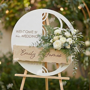 May include: A white wooden wedding sign with the text "WELCOME to our WEDDING" and the names "Emily & James" with the date 01.05.2026. The sign is decorated with white flowers and greenery and is displayed on a wooden easel.