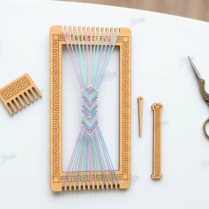 Op de afbeelding: Een houten weefgetouw met kleurrijke draden, een kleine kam en borduurgaren. Het weefgetouw heeft een geometrisch ontwerp en de tekst "THE AIS OF FATE". Ook inbegrepen zijn een naald, een decoratief houten stuk en een schaar.