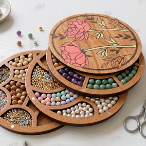 May include: Wooden bead organizer with multiple compartments filled with various beads in different colors and sizes. The top lid features a floral design with pink and green accents. A pair of scissors and tweezers are also visible.