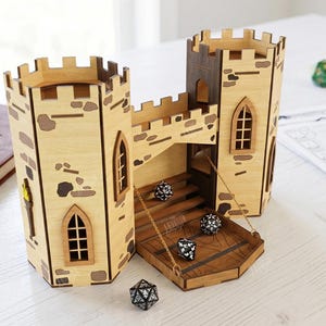 May include: A light-colored wooden dice tower featuring two castle turrets connected by a bridge with a drawbridge platform. The structure includes engraved stone details, arched windows, and a torch graphic. Three black dice are visible on the platform.