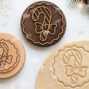 May include: Two circular wooden biscuit stamps featuring a candy cane with a bow design, one in dark wood and one in light wood, alongside a piece of rolled biscuit dough embossed with the same candy cane and bow pattern for baking.