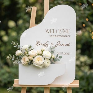 May include: A white wedding sign with a floral arrangement and the words "Welcome to the Wedding of Emily & James 01.05.2026". The sign is on a wooden easel. The sign is 3mm thick.
