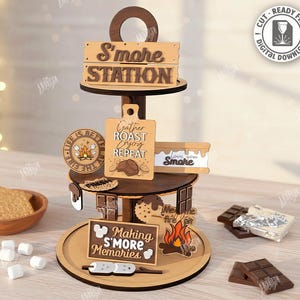 Może przedstawiać: Wielopoziomowa drewniana stacja s'mores z napisami "S'more Station" i "Making S'more Memories". Zawiera miskę z krakersami Grahama, piankami marshmallow i batonami czekoladowymi. Projekt jest wycinany laserowo i dostępny do pobrania cyfrowego.