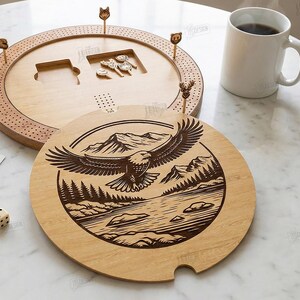 K&ouml;nnte beinhalten: Cribbage-Brett aus Holz mit rundem Deckel, der einen Adler &uuml;ber einer Berglandschaft zeigt. Das Brett enth&auml;lt Stifte und eine Spielfl&auml;che. Eine Tasse Kaffee, Spielkarten und W&uuml;rfel sind ebenfalls vorhanden. Der Text "CUT READY FILE DIGITAL DOWNLOAD" ist ebenfalls sichtbar.