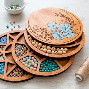 May include: Wooden bead organizer with multiple compartments filled with colorful beads. The top lid features a floral design with blue flowers. A spool of thread and a small bowl of beads are also present.