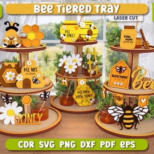 Può includere: Un vassoio a più livelli in legno a tema api. Il vassoio presenta tre livelli con elementi decorativi come favi, api e barattoli di miele. Il testo include "Bee Tiered Tray" e "Bee Sweet".