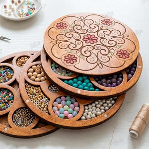 May include: Wooden bead organizer with multiple compartments filled with colorful beads. The top lid features a floral design. Other craft supplies, including scissors, tweezers, and thread, are also visible.
