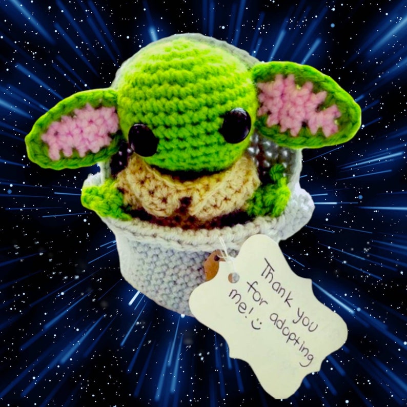 May include: A crocheted green and pink Baby Yoda plush toy sitting in a white crocheted pod. The toy has large black eyes and a small mouth. The pod has a white tag attached to it that says "Thank you for adopting me!"