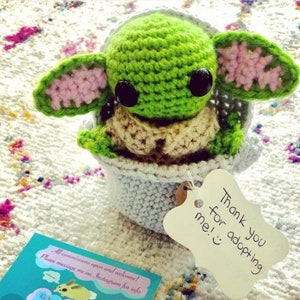 May include: A crocheted green and pink Yoda doll sitting in a blue crocheted pot. The doll has large black eyes and a small mouth. A white tag with the text "Thank you for adopting me!" is attached to the pot.