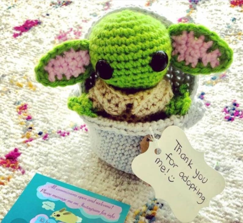 May include: A crocheted green and pink Yoda doll sitting in a blue crocheted pot. The doll has large black eyes and a small mouth. A white tag with the text "Thank you for adopting me!" is attached to the pot.