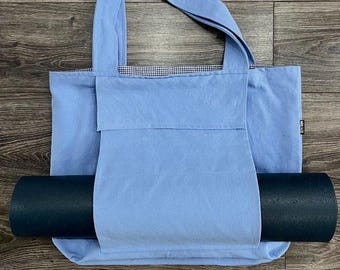 May include: A light blue canvas tote bag with a black yoga mat inside. The tote bag has a white and blue checkered lining and a pocket on the front.