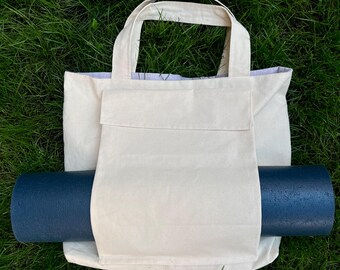 Op de afbeelding: Een witte canvas draagtas met een groot vak aan de voorkant. Een donkerblauwe yogamat is in de tas gestopt.