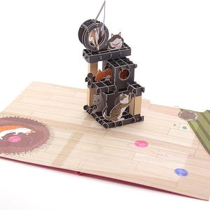 May include: A pop-up card depicting a black cat tree with multiple levels and several cat figures. The card is open, revealing a wooden floor design with a cat-shaped rug and colourful dots. A cat toy hangs from the top.