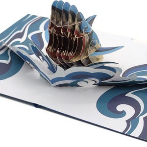 May include: A pop-up card depicting a 3D shark with open jaws, in blue and brown hues. The card's design includes a wave pattern in blue and white. The card is open, revealing the shark, and is set against a white background, evoking a sea theme.
