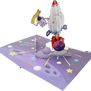 Puede incluir: Tarjeta de felicitación desplegable con un cohete blanco y rojo despegando hacia el espacio. Un astronauta flota cerca sosteniendo una pancarta que dice "CONGRAT!". La tarjeta tiene un fondo morado y azul con temática espacial con estrellas y planetas.