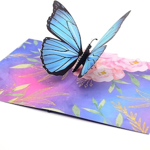 May include: A pop-up card with a blue butterfly and black detailing. The card has a watercolour design with pink flowers, green leaves, and gold accents on a purple and blue background.