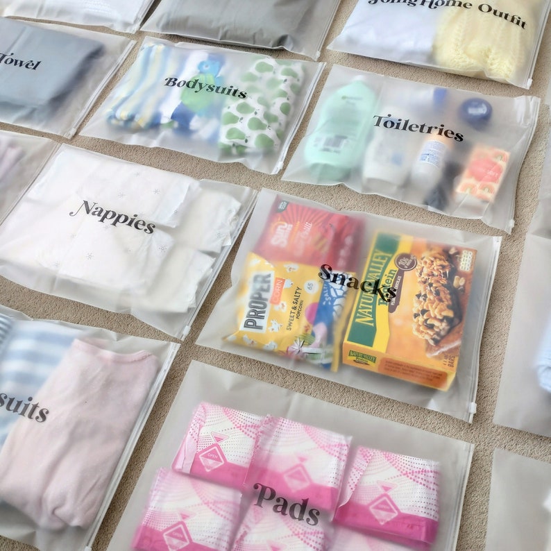 Może przedstawiać: Przezroczyste plastikowe torby z zamkiem błyskawicznym oznaczone r&oacute;żnymi kategoriami, takimi jak "Bodysuits", "Toiletries", "Nappies", "Snacks", "Pads" i "Towel". Torby są wypełnione r&oacute;żnymi przedmiotami, w tym ubraniami, artykułami toaletowymi, przekąskami i artykułami dla niemowląt.