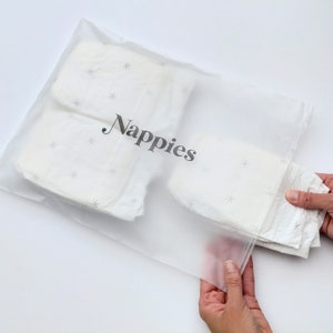 Może przedstawiać: Przezroczysty plastikowy worek z zamkiem błyskawicznym, oznaczony napisem "Nappies". Worek zawiera stos białych pieluch materiałowych z małym wzorem gwiazdek.