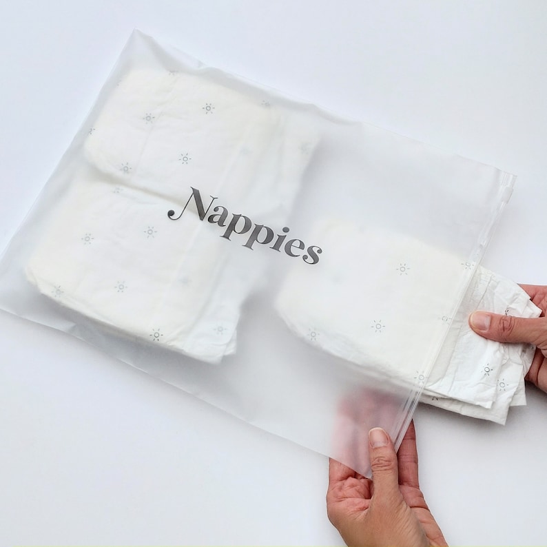 Może przedstawiać: Przezroczysty plastikowy worek z zamkiem błyskawicznym, oznaczony napisem "Nappies". Worek zawiera stos białych pieluch materiałowych z małym wzorem gwiazdek.