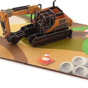 May include: A pop-up greeting card featuring a 3D excavator in orange and black. The card depicts a construction site with a traffic cone, barriers, and pipes. The card is open and displayed on a white surface.