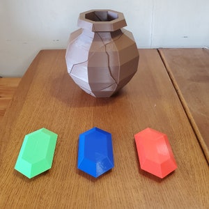 May include: Three 3D printed gems, one green, one blue, and one red, are arranged on a wooden surface. A brown, 3D printed, cracked pot sits above the gems.