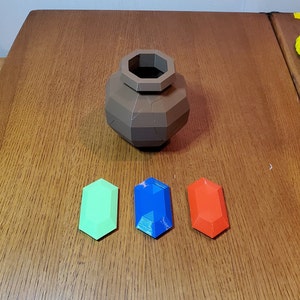 May include: Three 3D printed gems, one green, one blue, and one red, are arranged in a row on a wooden surface. A brown, 3D printed pot sits above the gems.