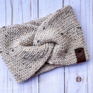 May include: A beige knitted headband with a twisted knot design. The headband has flecks of black throughout the yarn and a small brown leather tag with the text "Kennedy & Co". The headband is lying on a light wood surface.