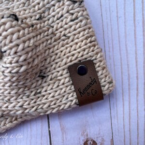 May include: Close-up of a beige knitted headband with black flecks. A brown leather tag with the text "Kennedy & Co" is attached. The headband is made of thick, textured yarn.