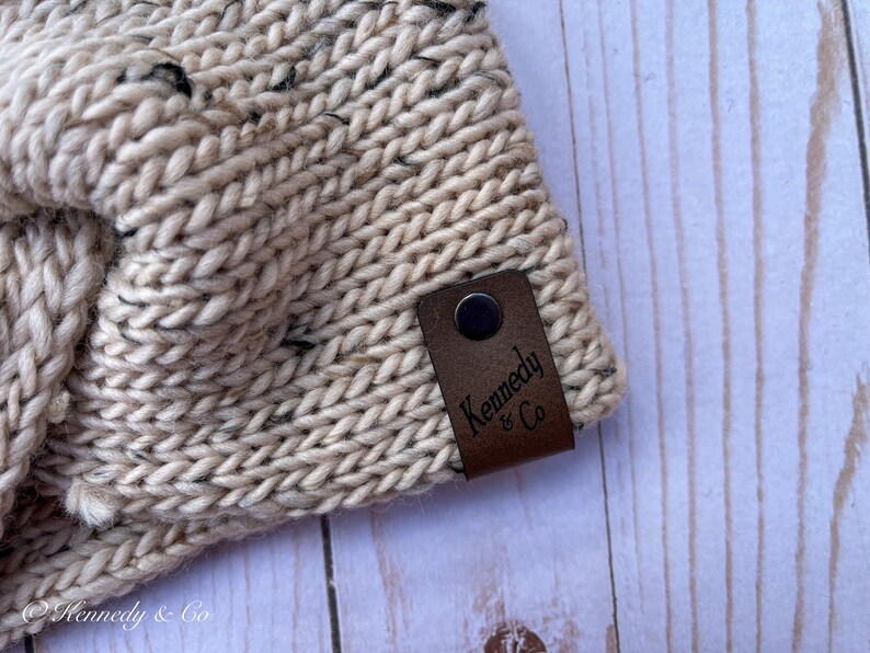 May include: Close-up of a beige knitted headband with black flecks. A brown leather tag with the text "Kennedy & Co" is attached. The headband is made of thick, textured yarn.
