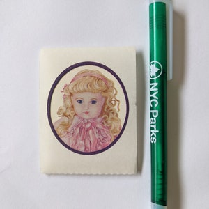 May include: A vintage sticker featuring a portrait of a doll with blonde curly hair, blue eyes, and a pink dress. The doll is framed by a purple circle. A green pen with "NYC Parks" printed on it is next to the sticker.