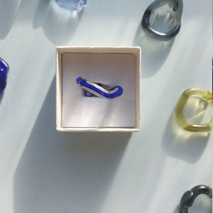 Op de afbeelding: Een blauwe glazen ring met een zilveren bandje ligt in een wit doosje. De ring is gevormd als een gebogen lijn. Het doosje staat op een lichtblauw oppervlak.