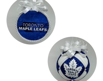 Pode incluir: Duas decorações de Natal. Uma é um círculo azul com o texto "TORONTO MAPLE LEAFS", uma borda de corda branca e um laço branco. A outra é uma bola azul e branca com um logotipo de folha de bordo e o mesmo texto.