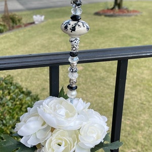 May include: A white ceramic flower pot with a black and white design and a beige fabric cover. The pot is decorated with a straw bow and contains white silk flowers. A crystal and bead embellishment is attached to the top of the pot.