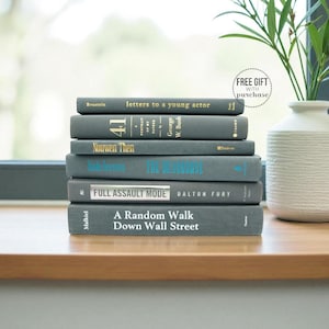 May include: A stack of six books with gray covers and various titles, including "Letters to a Young Actor" and "A Random Walk Down Wall Street." The books are arranged on a wooden surface near a window, with a white vase and greenery in the background.