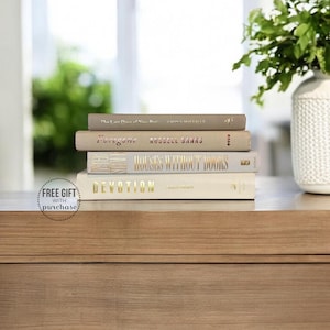 May include: A stack of four hardcover books with gold lettering on a wooden surface. The books are in neutral tones, with titles including "Foregone" and "Devotion". A white vase with greenery sits to the right. A sticker that says "FREE GIFT with purchase" is on the left.