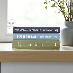 May include: Three hardcover books stacked on a wooden surface. The top book is navy blue and reads "THE WOMAN IN CABIN 10". The middle book is light blue and reads "BEFORE THE FALL". The bottom book is green and reads "THE SEARCH". A vase with greenery is in the background.