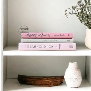 May include: A white bookshelf with three pink books stacked on the top shelf. The top book is titled "Raising the Barre" by Lauren Kerster. The middle book is titled "Best Friends Forever" by  Nimbeila Lawson Roby. The bottom book is titled "The Last Collection..." by Jeanne Mackin. A small wooden bowl with dried flowers sits on the top shelf. A white crocheted vase sits on the bottom shelf with a wooden branch in front of it.