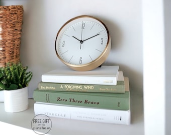 May include: A gold clock with black hands and Roman numerals sits on a stack of books. The books are titled "A Forgiving Wind", "Zero Three Bravo", and "Struggle".
