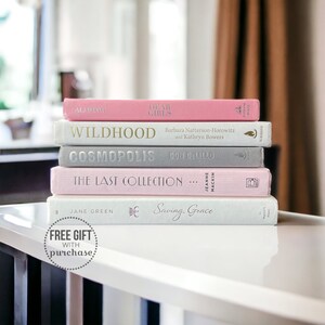 K&ouml;nnte beinhalten: Ein Stapel von f&uuml;nf Hardcover-B&uuml;chern in Rosa-, Grau- und Wei&szlig;t&ouml;nen. Die Titel und Autoren sind sichtbar, darunter "Dear Girls" und "Wildhood". Die B&uuml;cher sind auf einer wei&szlig;en Oberfl&auml;che gestapelt, mit einer Nachricht "Free Gift With Purchase".