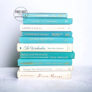 May include: A stack of hardcover books in various colors, including teal and white. The book titles and author names are visible, with gold lettering. The books are arranged in a neat pile, creating a visually appealing display.