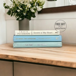 May include: A stack of three books on a light wood surface. The top book is white with gold lettering and the title "Secrets of My Heart" by Tracie Peterson. The middle book is light blue with gold lettering and the title "Between Two Kingdoms" by Suleika Jaouad. The bottom book is a lighter shade of blue with gold lettering and the title "Plum Spooky" by Jumel Exant. A white sticker with the text "FREE GIFT WITH purchase" is visible in the upper right corner of the image.