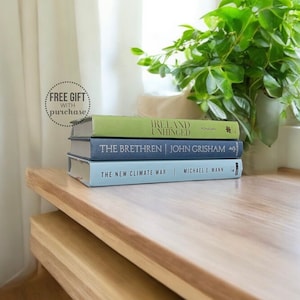 May include: Three hardcover books stacked on a wooden surface. The top book is green and titled "Ireland Unhinged" by Monaghan. The middle book is blue and titled "The Brethren" by John Grisham. The bottom book is light blue and titled "The New Climate War" by Michael E. Mann.