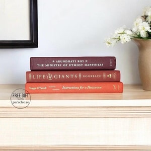 May include: Three hardcover books stacked on a light wood shelf. The top book is maroon with white text that reads "Arundhati Roy * The Ministry of Utmost Happiness". The middle book is red with white text that reads "Life/Giants Roobach". The bottom book is orange with white text that reads "Maggie O'Farrell Instructions for a Heatwave".