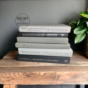 May include: A stack of seven books with varying shades of gray and black spines. Titles include "The Ungarnished Truth," "Disciplines of the Heart," and "Daughters of the Storm." The books are arranged on a wooden surface.