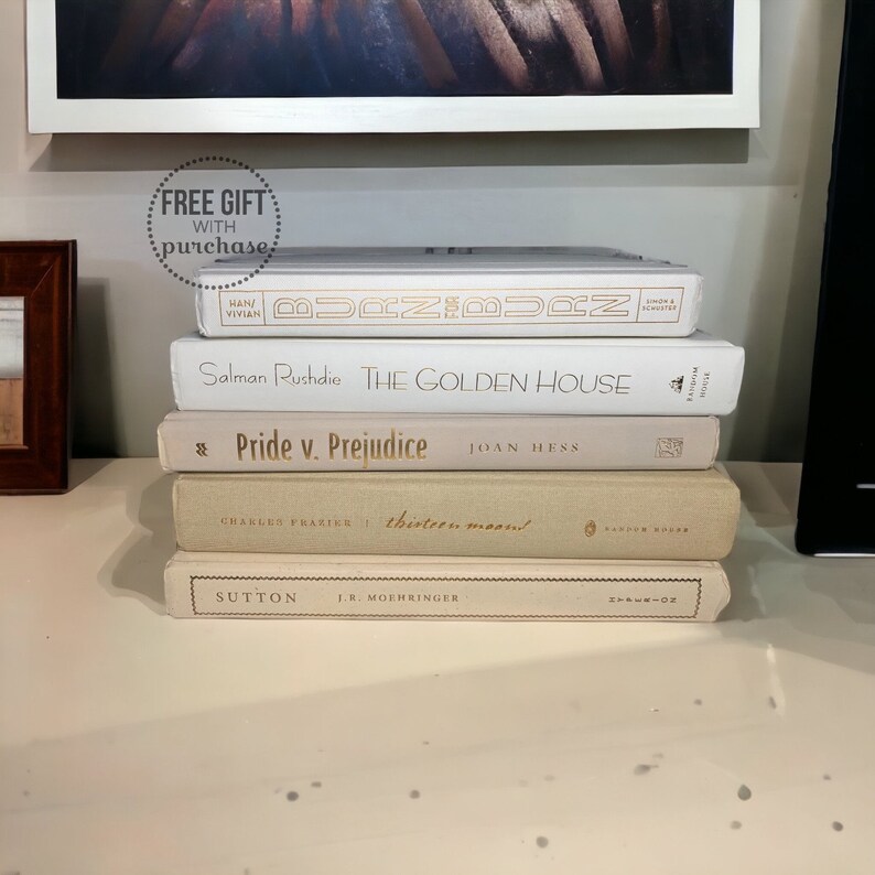 May include: A stack of five books with white covers and gold lettering. The titles are "Burn for Burn", "The Golden House", "Pride v. Prejudice", "thirteen moons!" and "Sutton".