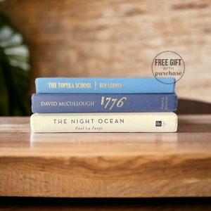 May include: Three hardcover books stacked on a wooden surface. The top book is light blue with the title "THE TOPEKA SCHOOL" and author "BEN LERNER". The middle book is navy blue with "1776" in gold lettering. The bottom book is off-white with the title "THE NIGHT OCEAN".