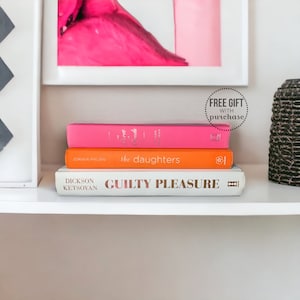 May include: A shelf displays three books with colorful covers. The top book is pink, the middle is orange, and the bottom is white with gold lettering that reads "GUILTY PLEASURE." A framed pink artwork hangs above.