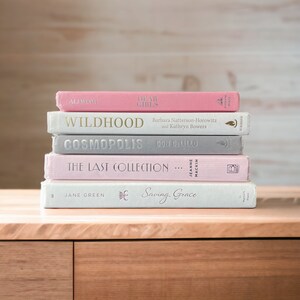 K&ouml;nnte beinhalten: Ein Stapel gebundener B&uuml;cher in Rosa, Grau und Wei&szlig;. Titel sind "Dear Girls", "Wildhood", "Cosmopolis", "The Last Collection" und "Saving Grace". Die B&uuml;cher liegen auf einer Holzoberfl&auml;che vor einem hellen Holz Hintergrund.