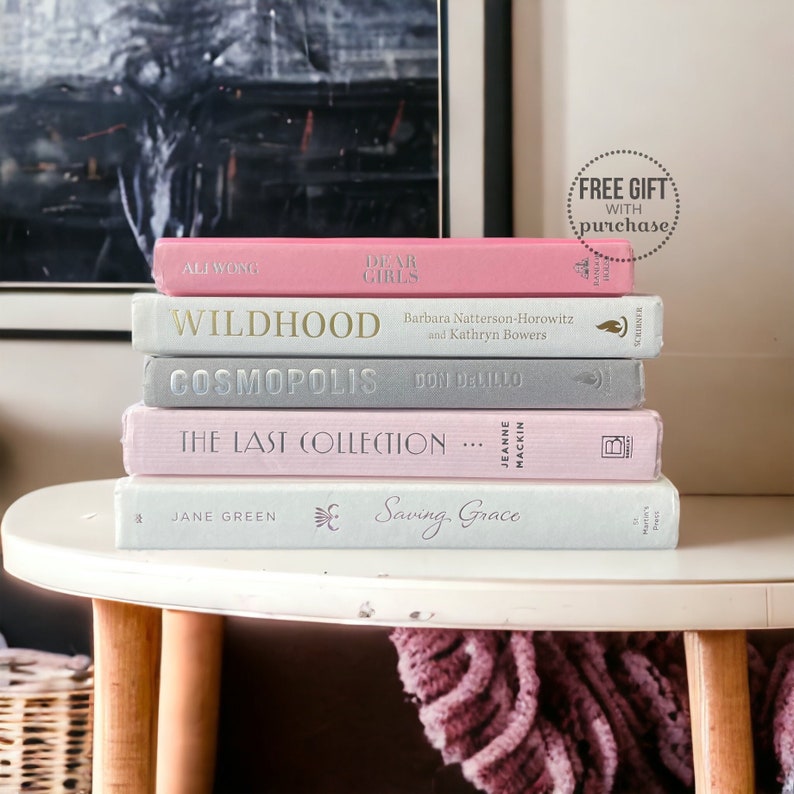 K&ouml;nnte beinhalten: Ein Stapel gebundener B&uuml;cher in Pastellfarben, darunter Rosa und Grau, auf einem kleinen, runden wei&szlig;en Tisch. Titel wie "Dear Girls" und "Cosmopolis" sind sichtbar. Ein Aufkleber im Hintergrund lautet "Free Gift with Purchase".