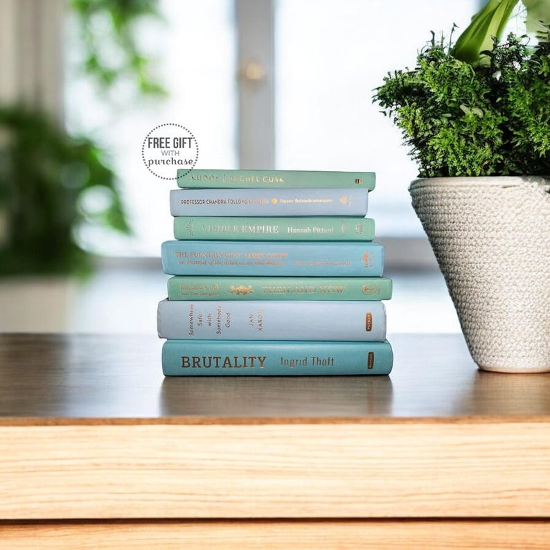 May include: A stack of seven light blue hardcover books with gold lettering on a wooden surface. The books are titled "A Long Petal of the Sea", "Professor Chandra Follows His Nose", "Vigdis Empire", "The Unlikely Pilgrimage of Harold Fry", "Some of Us Are Breathing", "The God of Small Things", and "BRUTALITY". A potted green plant is in the background.